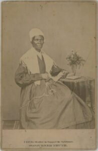 A historical sepia toned photo portrait of an older African American woman sitting beside a small round table, with a work of knitting in her lap, dressed in period clothing. A quote at the bottom of the photo: "I Sell the Shadow to Support the Substance".Nineteenth century.